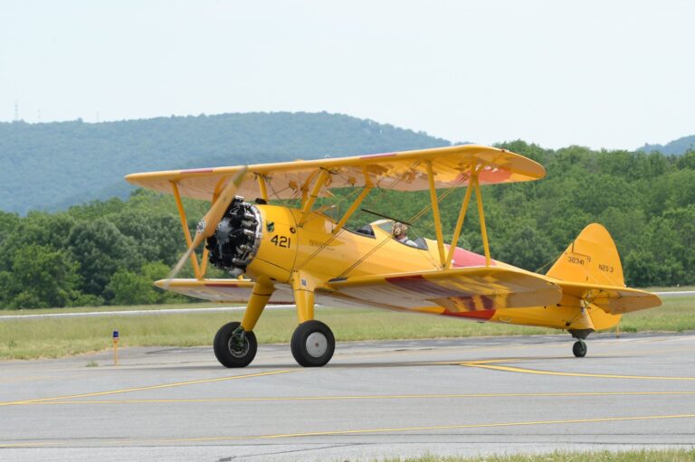 Yellow Boeing Stearman on tarmac with pilot, ready for takeoff, surrounded by nature.